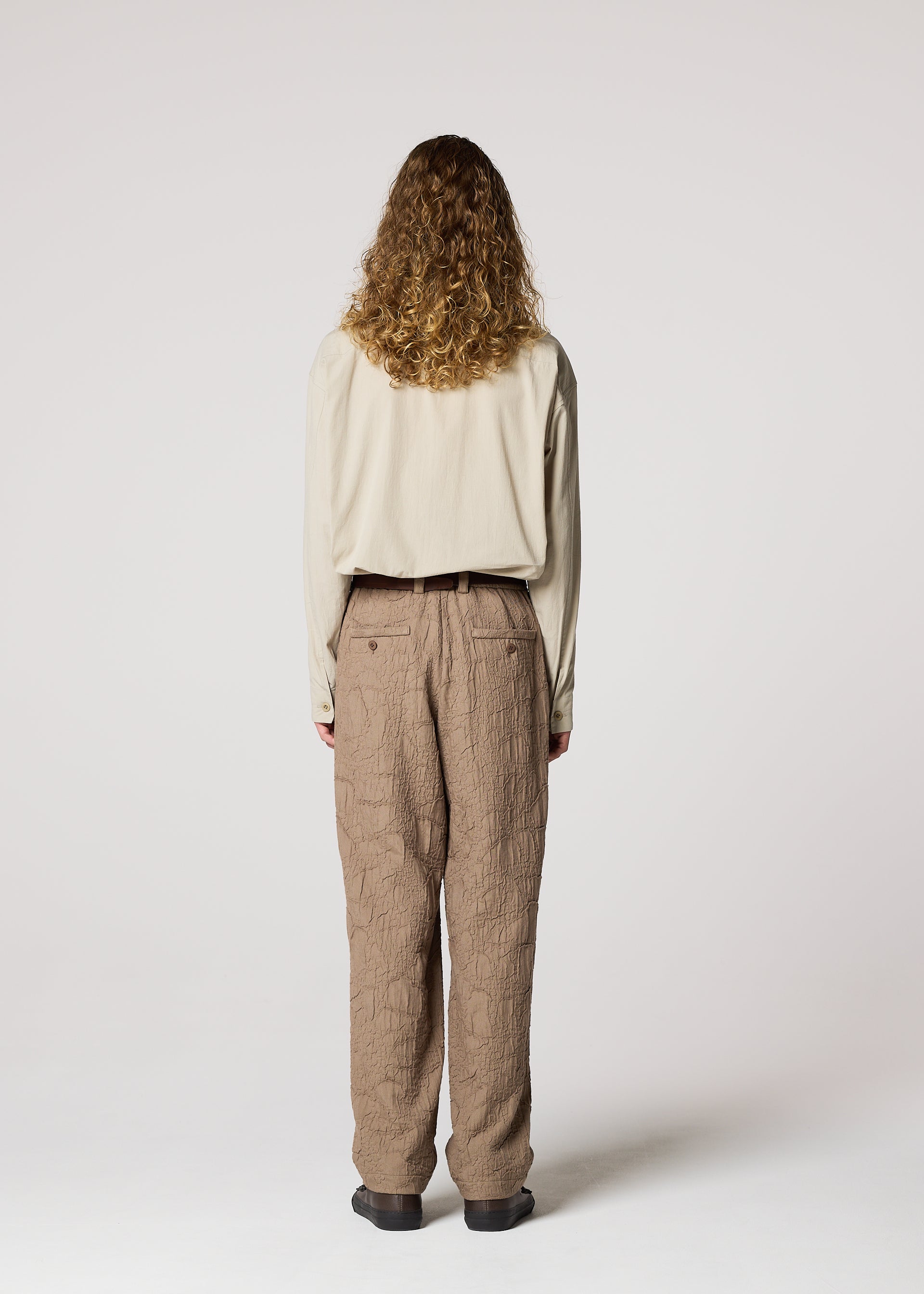 Zoomed view A person with long curly hair faces away, wearing the IM MEN SHRINK JACQUARD beige long-sleeve shirt and loose brown wood-grain woven pants with unique texture, set against a plain light background.