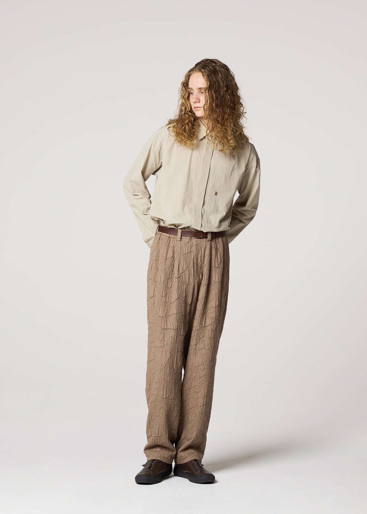 A person with long curly hair stands against a light background, wearing IM MEN's SHRINK JACQUARD beige button-up shirt and loose textured wood-grain woven pants, paired with a brown belt and dark shoes, hands behind their back. view 8 of 8