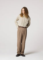 A person with long curly hair stands against a light background, wearing IM MEN's SHRINK JACQUARD beige button-up shirt and loose textured wood-grain woven pants, paired with a brown belt and dark shoes, hands behind their back.