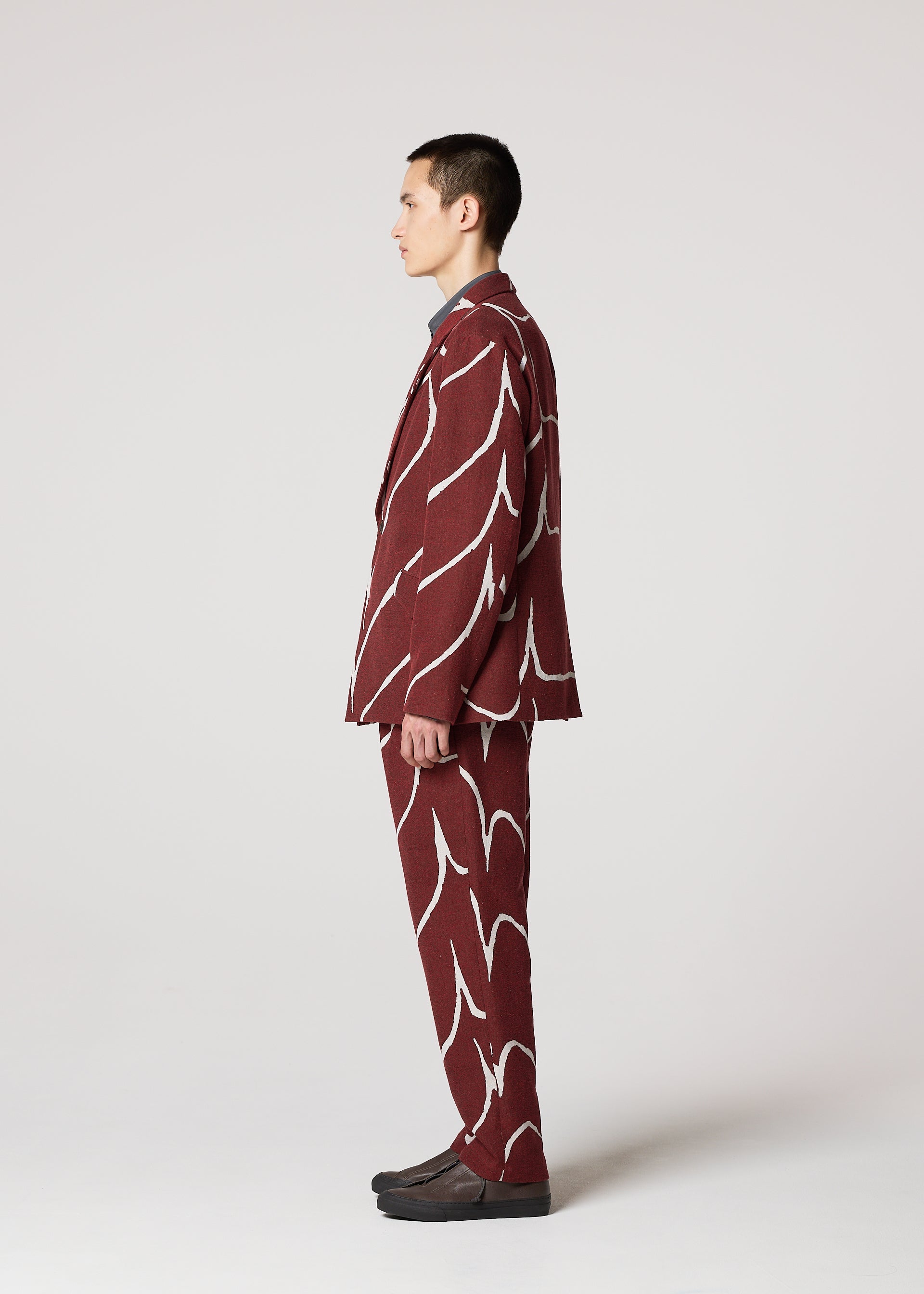 Zoomed view A person with short hair stands in profile against a plain background, wearing IM MEN's EARTH maroon suit featuring bold wave-pattern pants and dark shoes.