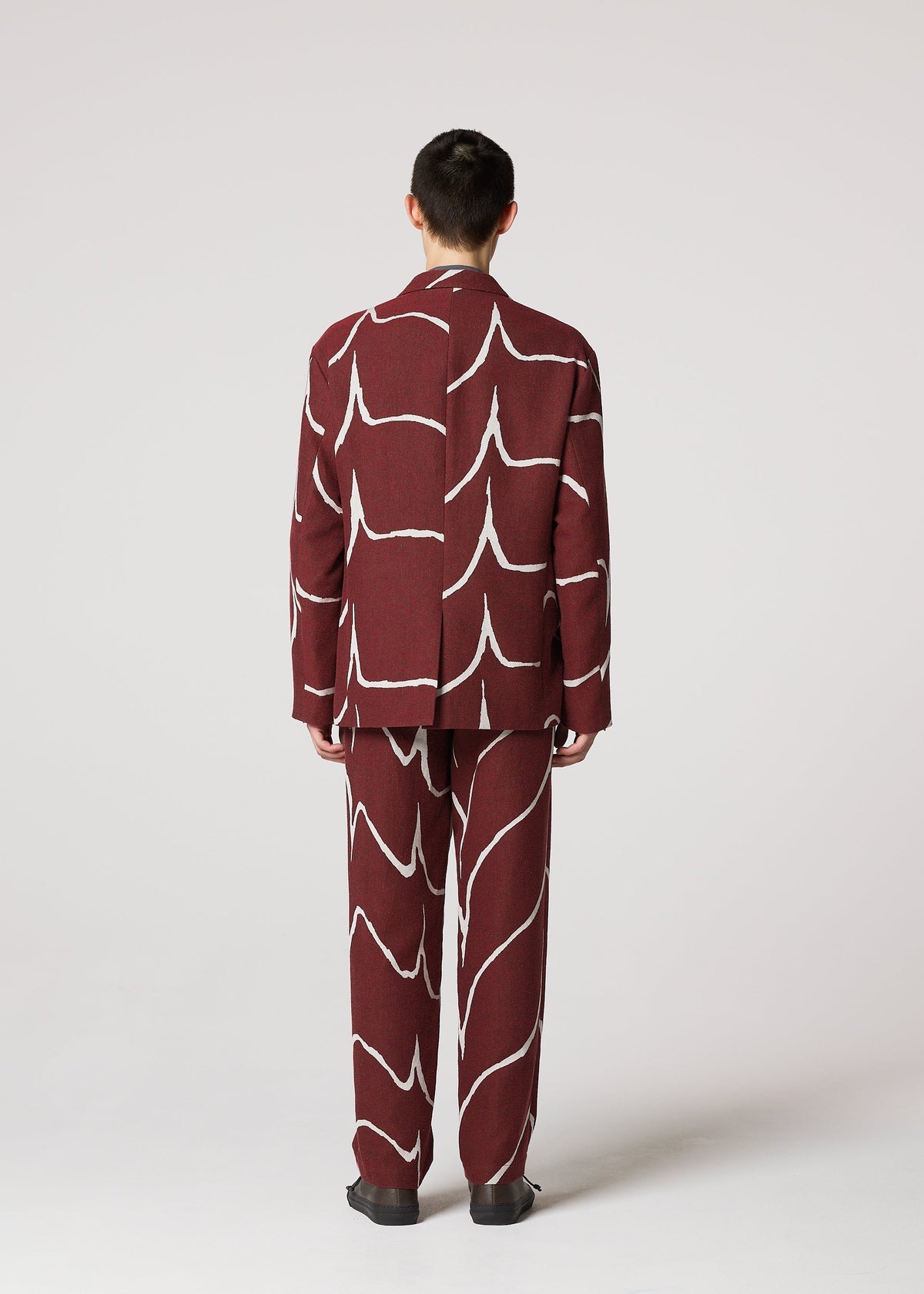 A person faces away, wearing the IM MEN EARTH suit—a maroon long jacket and matching trousers with bold white abstract lines and wave patterns—set against a plain light gray background. view 7 of 8