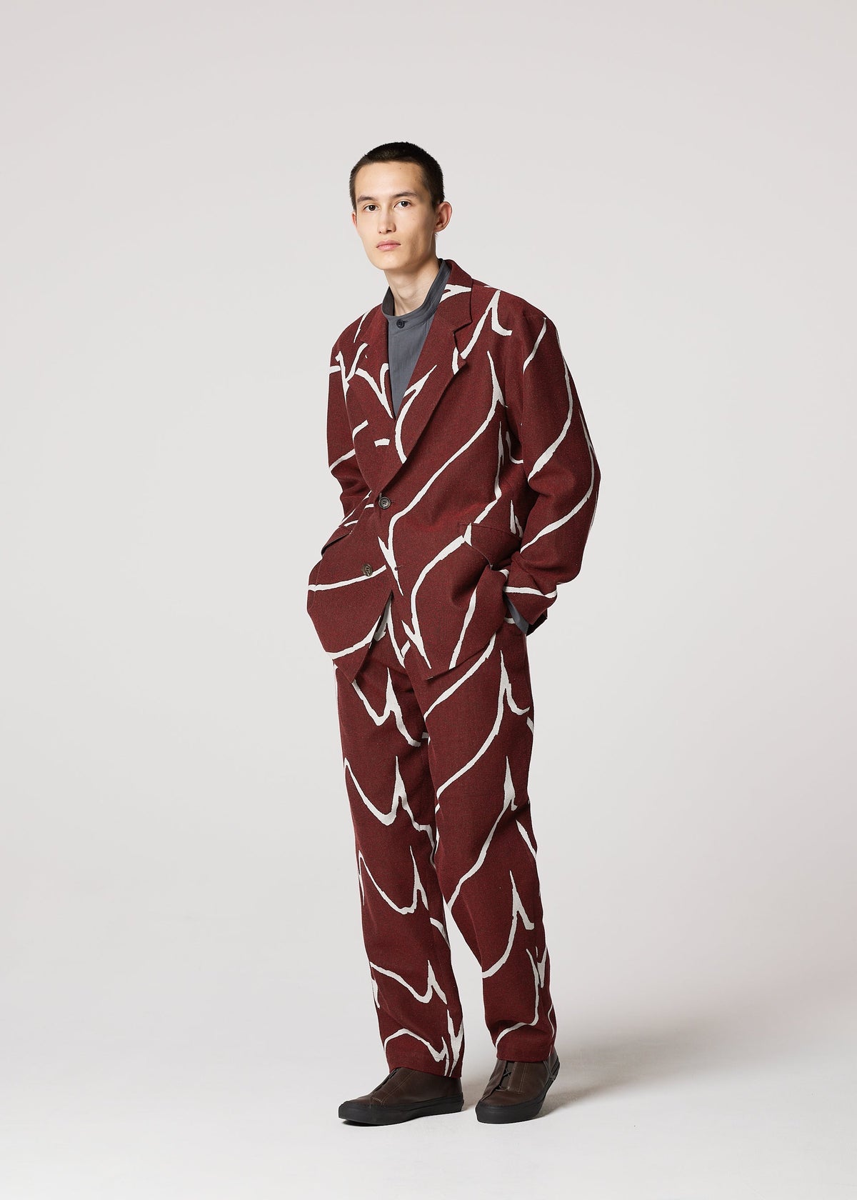 Against a plain light background, a person wears IM MEN's EARTH suit: a loose maroon set with bold ceramic-inspired white abstract patterned pants, paired with a gray shirt and dark shoes. Hands in pockets, expression neutral. view 8 of 8