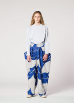 A person with long curly hair stands facing forward in a loose white long-sleeve shirt and IM MEN's INLAY wide, high-waisted jacquard loom pants with a bold blue and gray wave pattern, set against a plain light background.