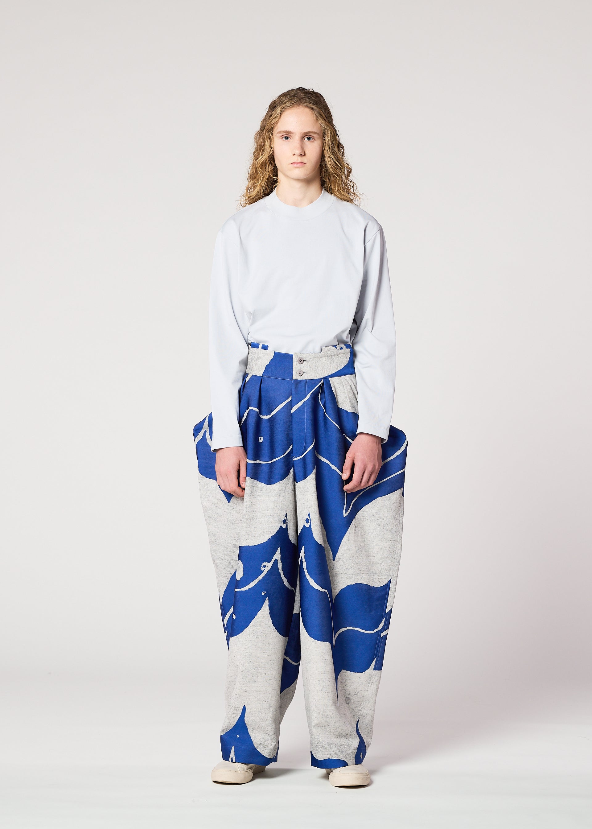 Zoomed view A person with long curly hair stands facing forward in a loose white long-sleeve shirt and IM MEN's INLAY wide, high-waisted jacquard loom pants with a bold blue and gray wave pattern, set against a plain light background.