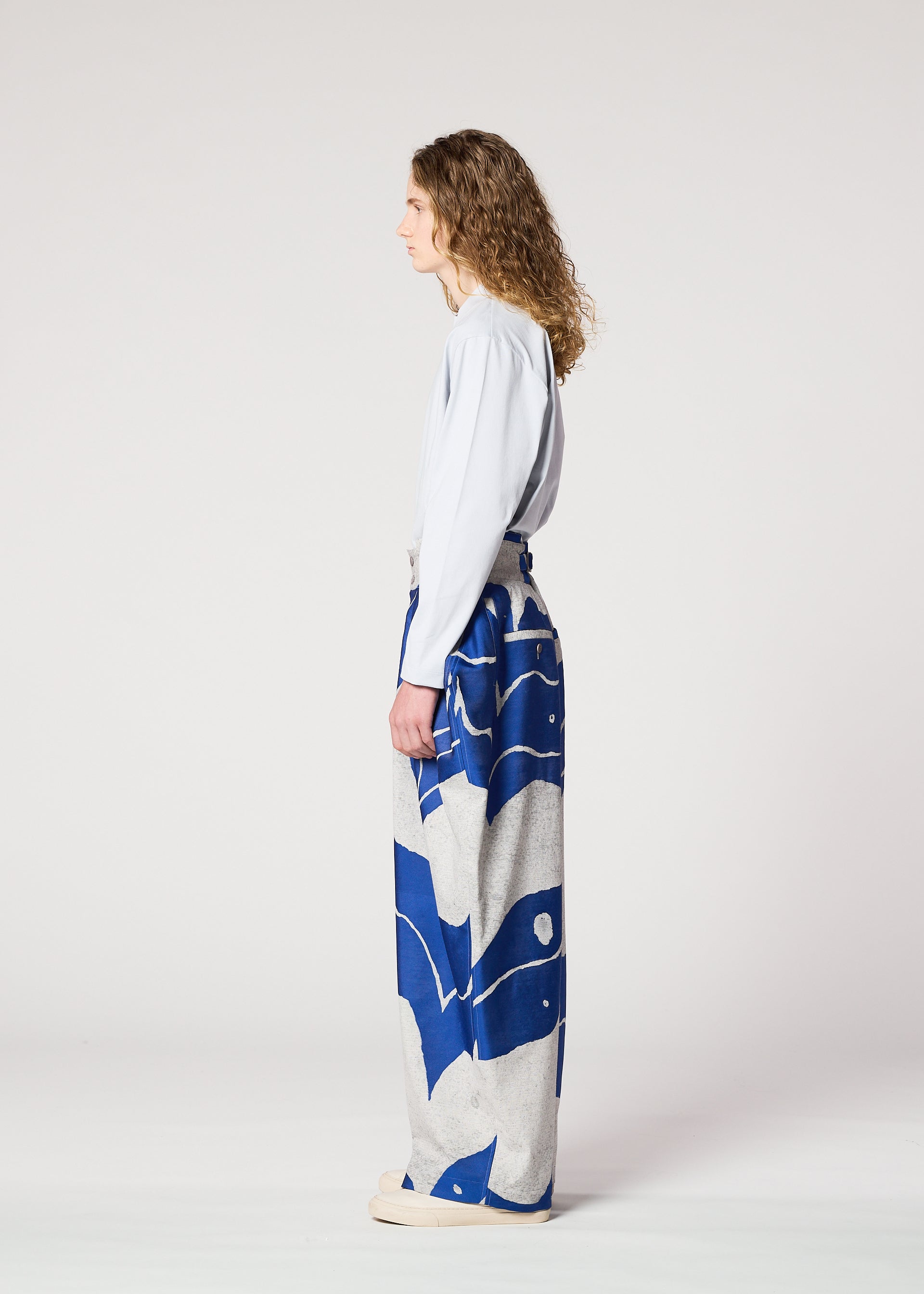 Zoomed view A person with long curly hair stands in profile against a plain background, wearing IM MEN's INLAY: a light blue long-sleeved top and high-waisted, wide-legged jacquard loom pants with blue and gray patterns, paired with cream shoes.