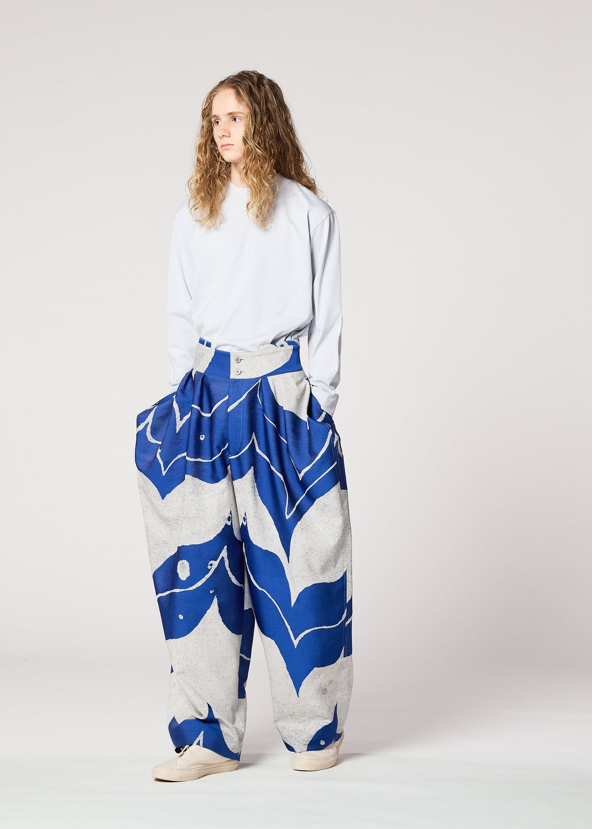 Wearing IM MEN’s INLAY, a loose white long-sleeve shirt with oversized ceramic-inspired pants featuring bold blue abstract patterns, a person with long curly hair stands against a plain light background. view 6 of 6