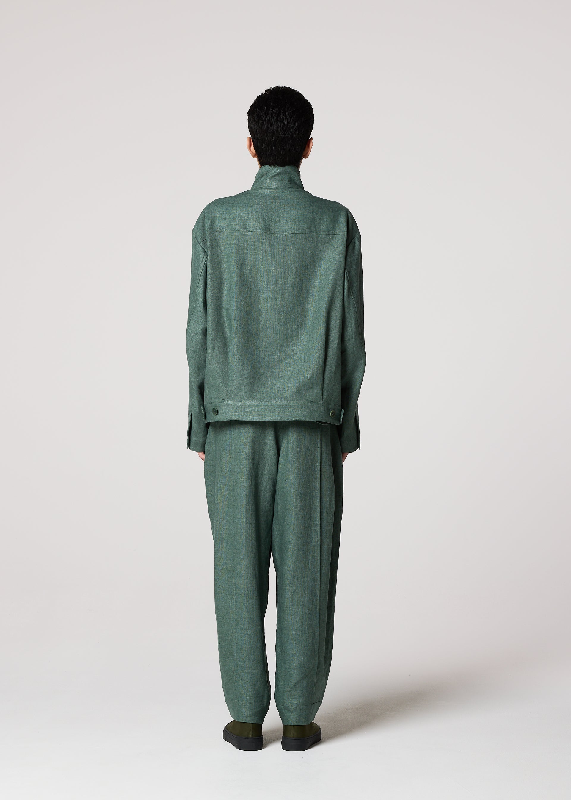 Zoomed view A person faces away from the camera, wearing IM MEN's TOUGH LINEN suit in green. The loose-fitting design features long sleeves and durable linen pants with a natural texture, set against a plain, light-colored background.