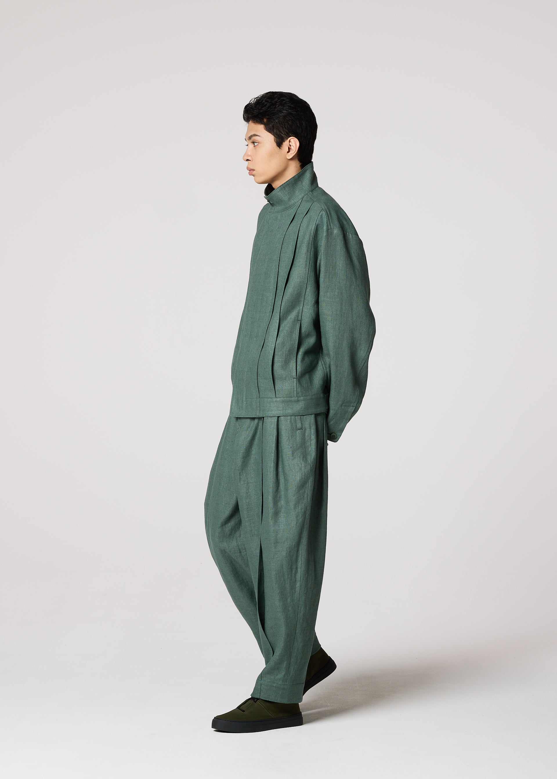 Zoomed view A person stands in profile against a plain light background, wearing IM MEN’s TOUGH LINEN: a loose-fitting, matching green jacket and durable linen pants with natural texture, hands behind their back and dark slip-on shoes.