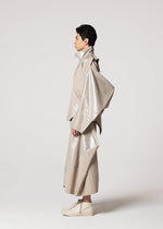 A person stands in profile wearing IM MEN's avant-garde GINTO FLAT set with glossy, asymmetrical panels and plant-based polyester pants, paired with matching shoes, all in light beige against a plain background.