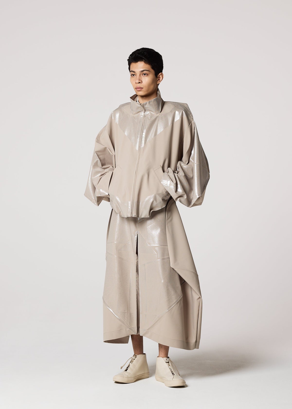 A person stands against a plain background wearing IM MEN's GINTO FLAT, an oversized shiny beige jacket with wide sleeves, a matching long skirt, light high-tops, and ceramic-inspired pants for an artistic touch. view 9 of 9