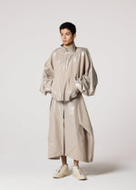 A person stands against a plain background wearing IM MEN's GINTO FLAT, an oversized shiny beige jacket with wide sleeves, a matching long skirt, light high-tops, and ceramic-inspired pants for an artistic touch.