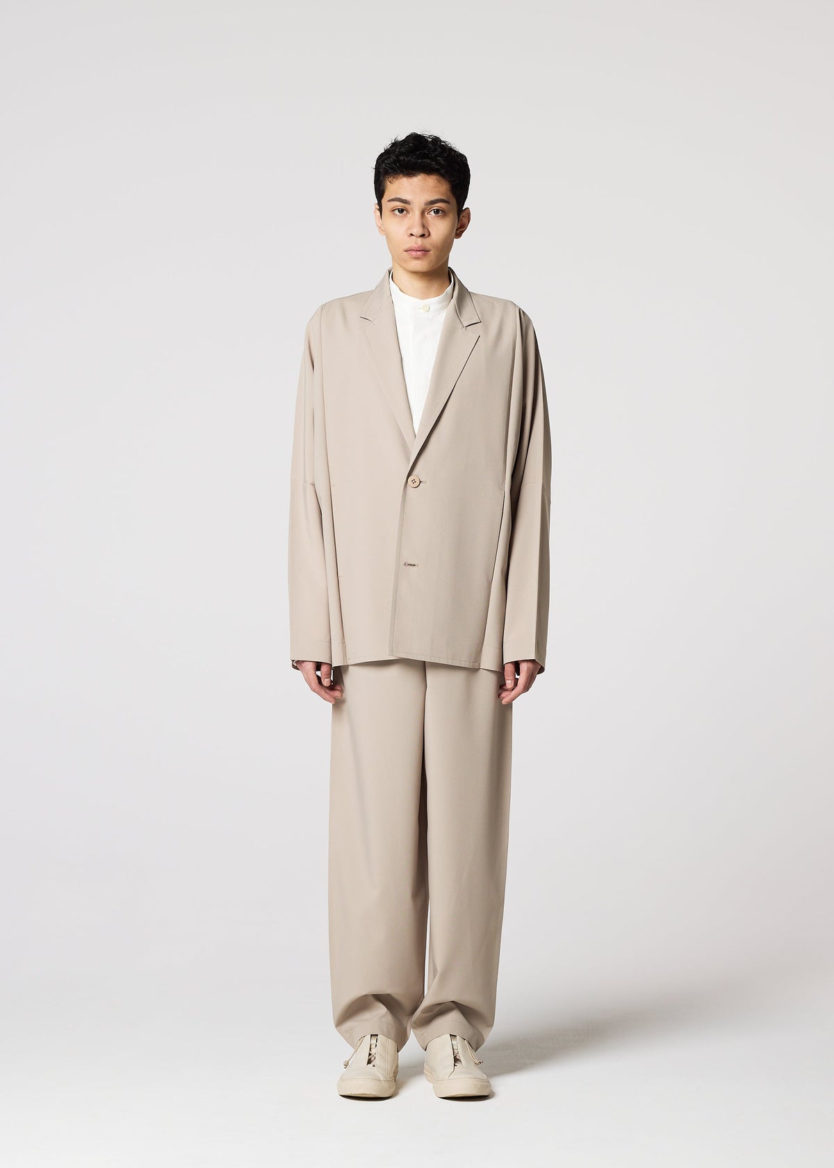 A person stands facing forward, wearing the IM MEN FLAT oversized beige suit with wrinkle-resistant pants, a white shirt, and beige sneakers against a plain light background. view 5 of 8