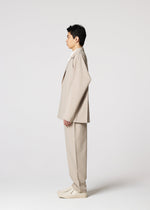 A person stands in profile facing left, wearing the IM MEN FLAT oversized beige suit with wrinkle-resistant pants and matching shoes, against a plain white background.