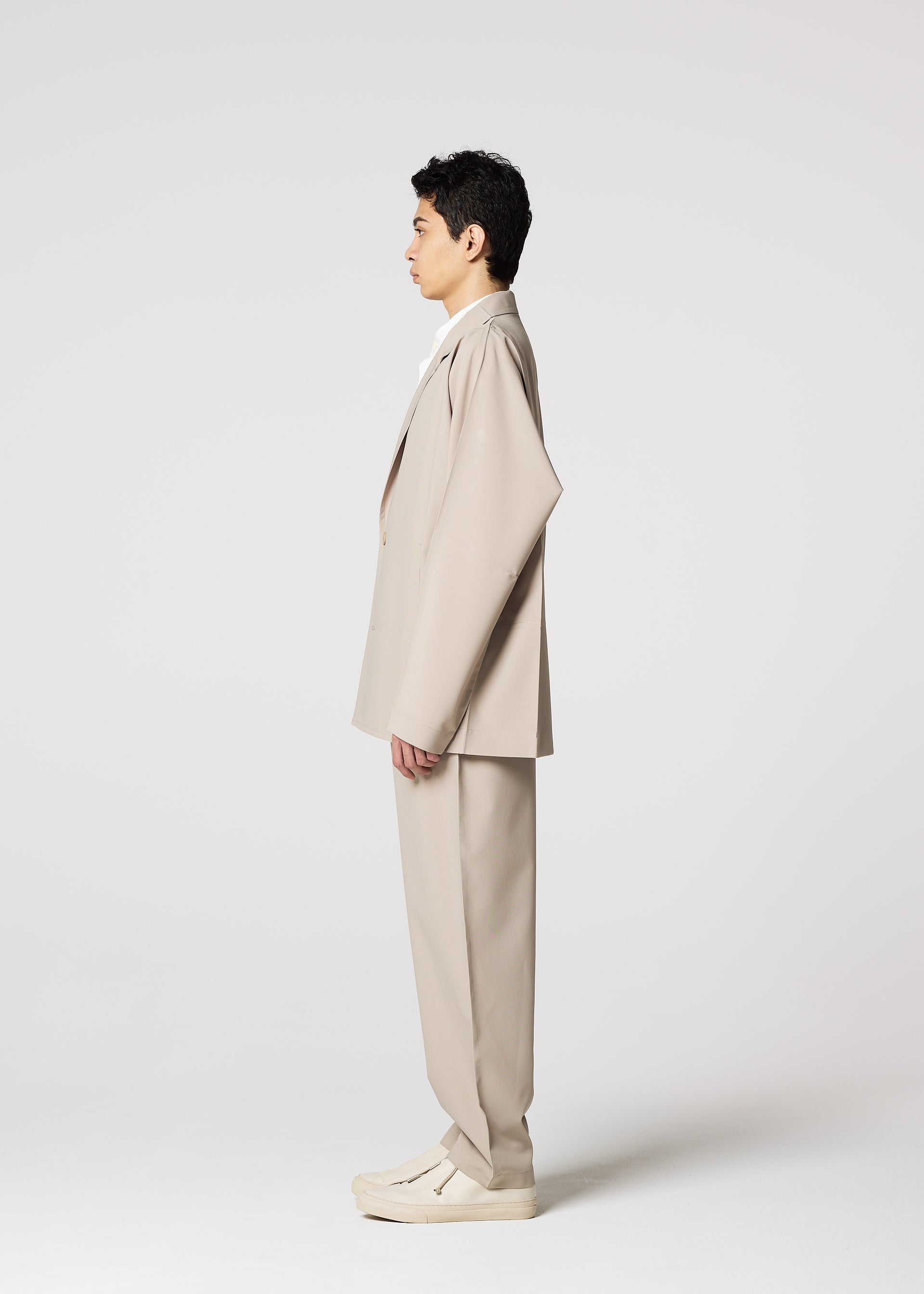Zoomed view A person stands in profile facing left, wearing the IM MEN FLAT oversized beige suit with wrinkle-resistant pants and matching shoes, against a plain white background.