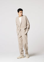 A man in IM MEN’s FLAT wrinkle-resistant pants, styled as part of a loose beige suit with a white t-shirt and beige shoes, stands with hands in pockets against a plain light background, looking to the side.