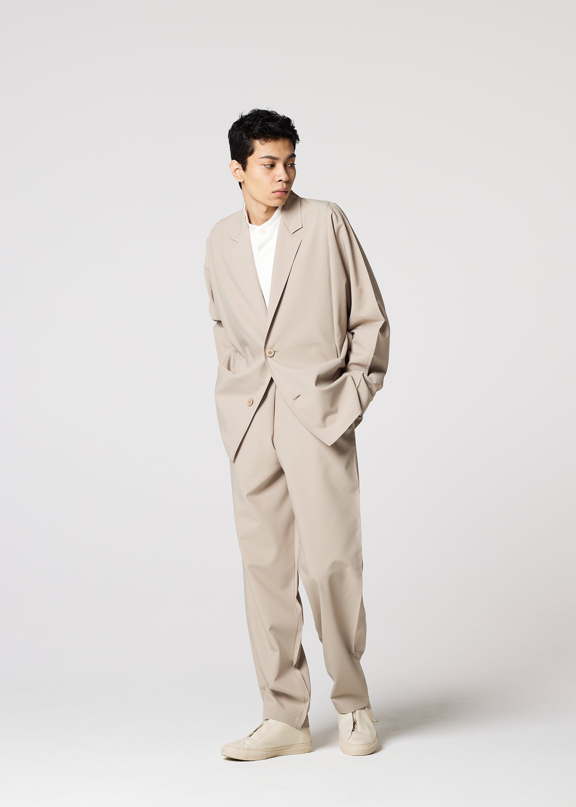 Zoomed view A man in IM MEN’s FLAT wrinkle-resistant pants, styled as part of a loose beige suit with a white t-shirt and beige shoes, stands with hands in pockets against a plain light background, looking to the side.