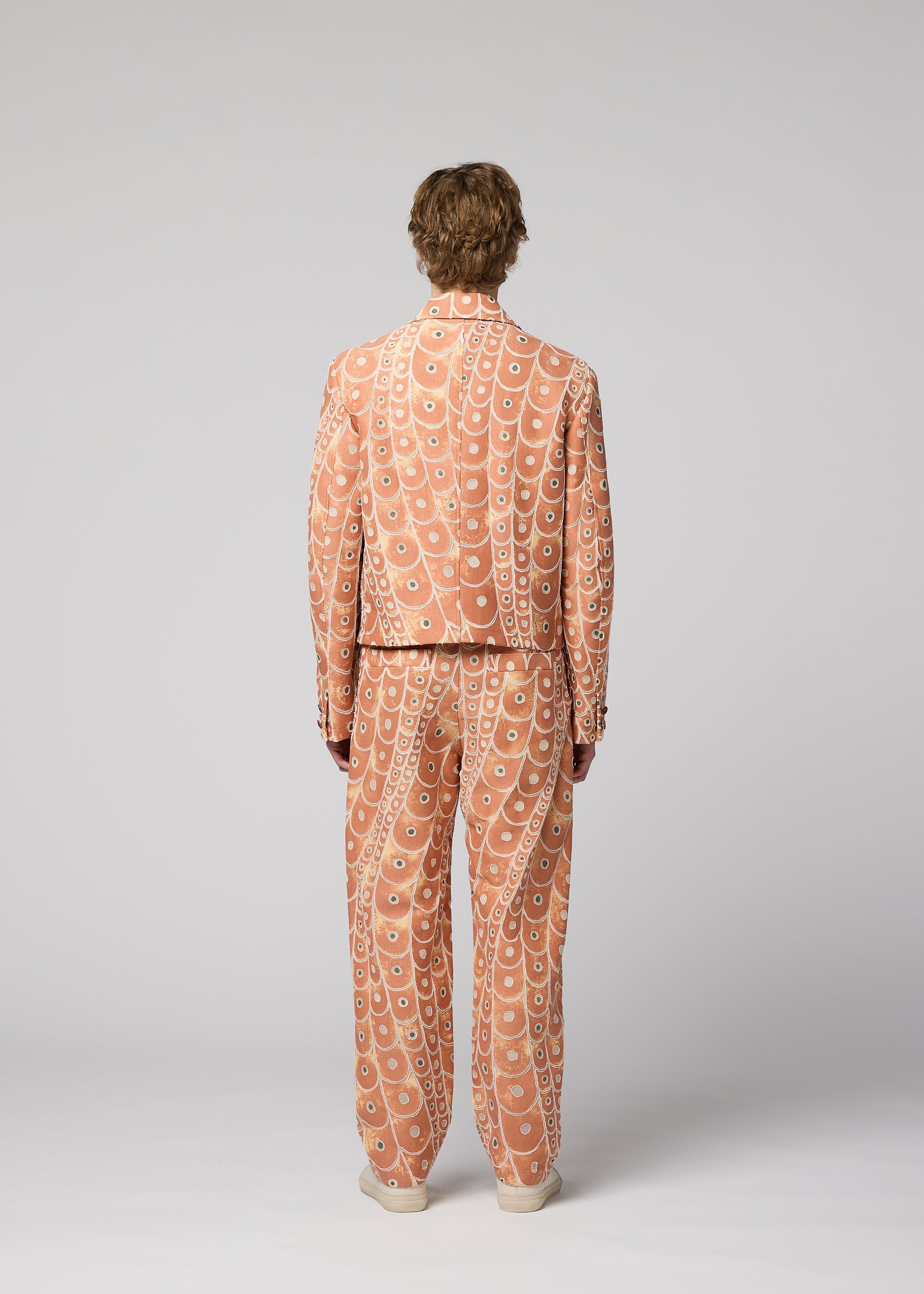 Zoomed view A person faces away, wearing the IM MEN UROKOMON suit in peach with scale-like designs and a wavy circular white pattern, set against a plain light gray background.