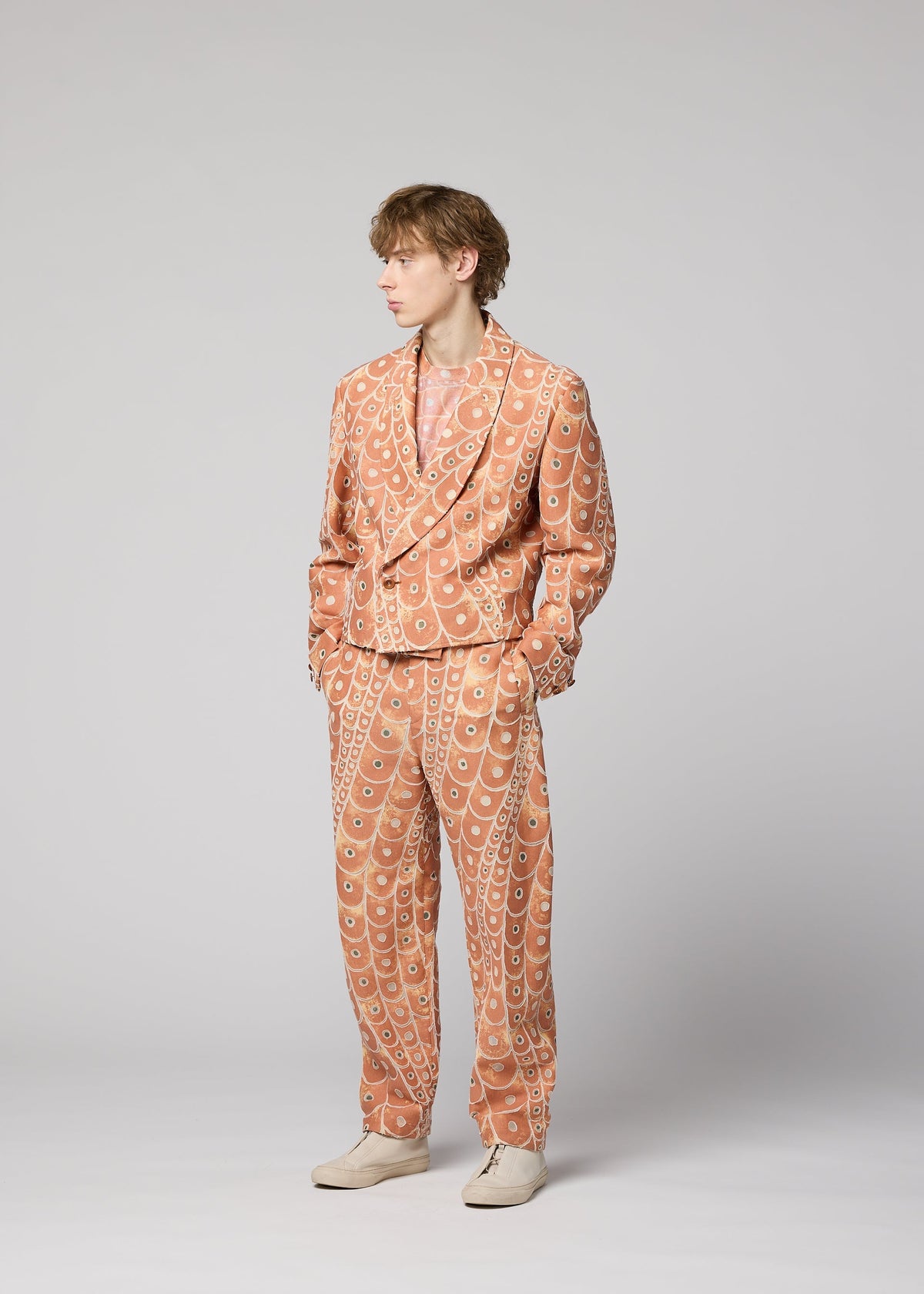 A person stands against a light gray background, wearing the IM MEN UROKOMON peach suit with abstract white, scale-like designs and a matching shirt, paired with cream shoes. The person looks to the side. view 5 of 7