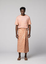 A man faces forward in IM MEN’s UROKOMON: a peach short-sleeve shirt and textured wrap skirt with a white geometric pattern inspired by Shoji Kamoda, paired with brown sandals. The background is plain light gray.