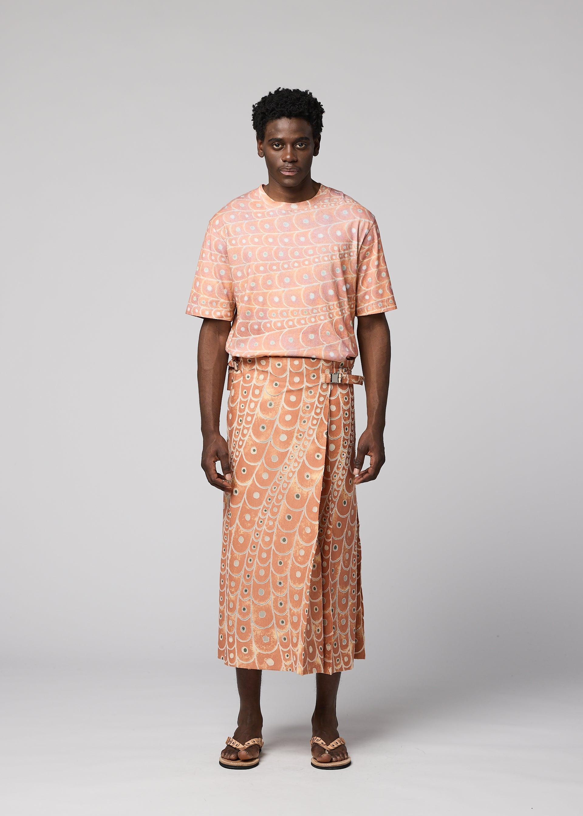 Zoomed view A man faces forward in IM MEN’s UROKOMON: a peach short-sleeve shirt and textured wrap skirt with a white geometric pattern inspired by Shoji Kamoda, paired with brown sandals. The background is plain light gray.