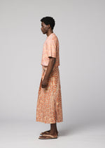 A person stands in profile against a light gray background, wearing IM MEN's UROKOMON: a peach short-sleeve top and a white and orange circular-patterned textured wrap skirt, paired with sandals.