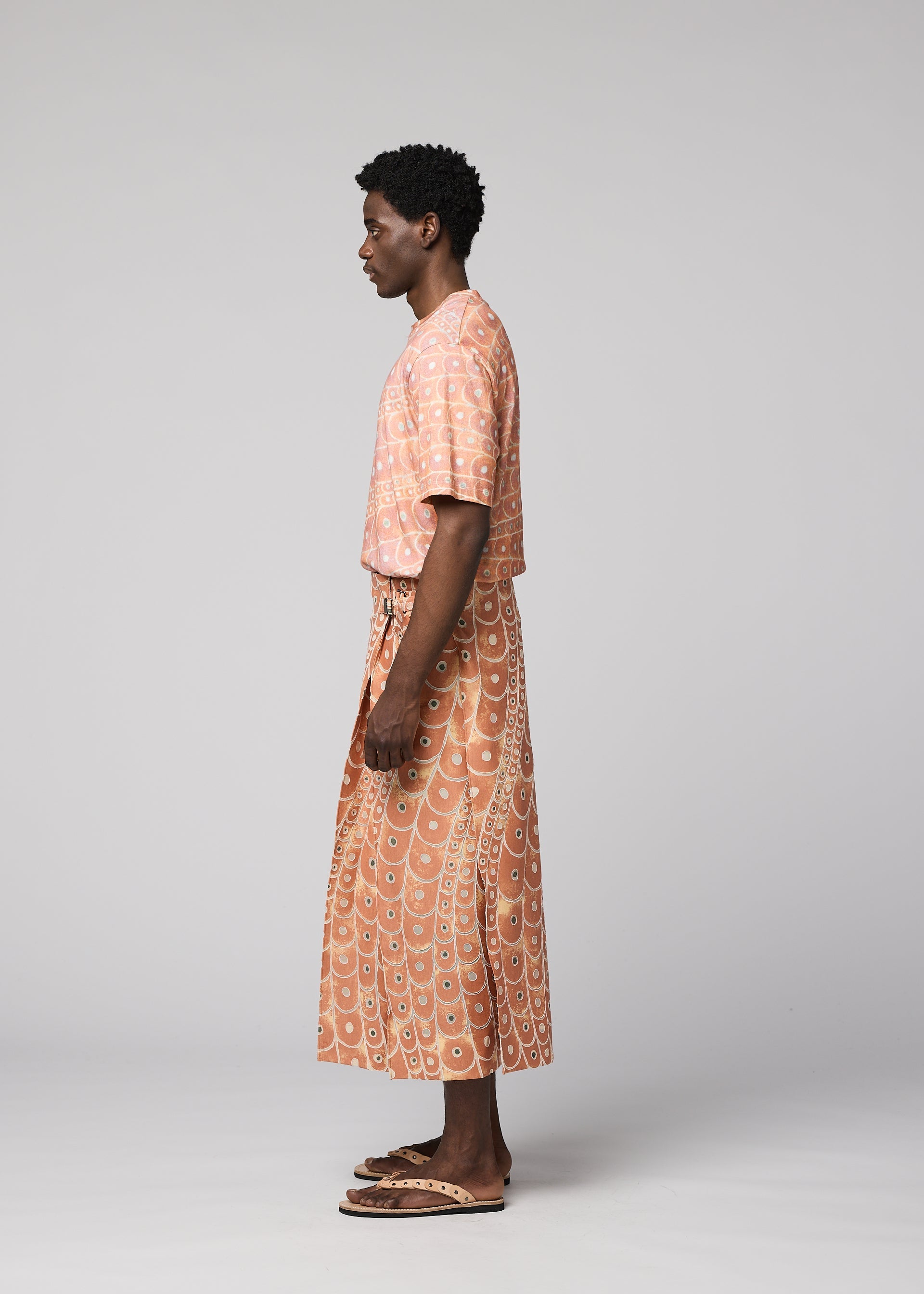 Zoomed view A person stands in profile against a light gray background, wearing IM MEN's UROKOMON: a peach short-sleeve top and a white and orange circular-patterned textured wrap skirt, paired with sandals.