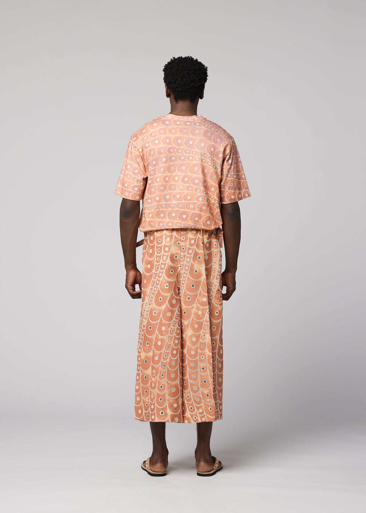 A person faces away, wearing the IM MEN UROKOMON set—a peach top and wide-legged pants with a white geometric pattern—paired with sandals, against a plain light gray background. view 5 of 6