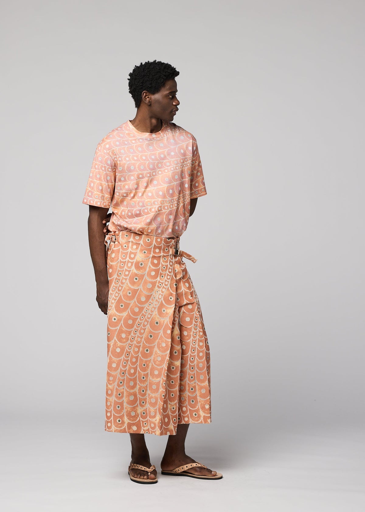 A person models the IM MEN UROKOMON set—matching peach pants and a short-sleeve shirt with white circular patterns, inspired by ceramics. Sandals complete the look as the person gazes to the side against a plain background. view 6 of 6