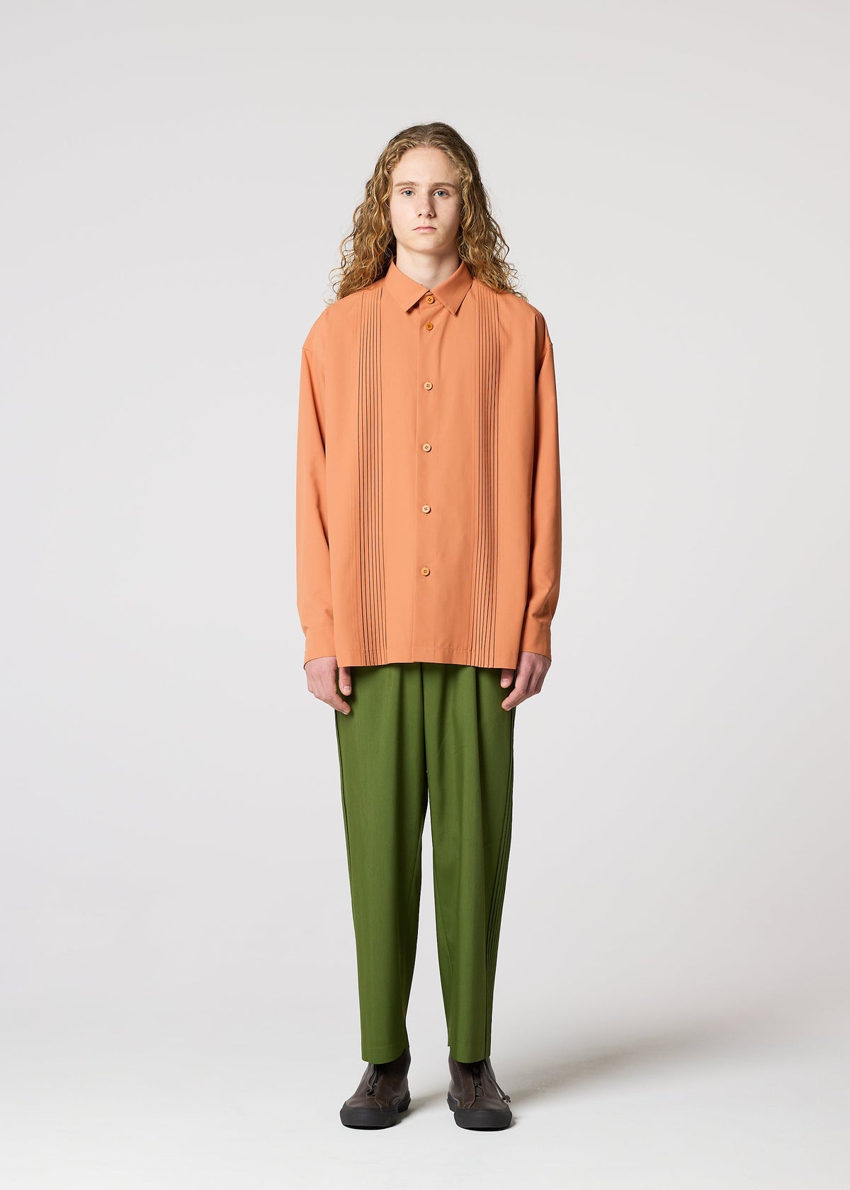 A person with long curly hair stands facing forward, wearing IM MEN's AIR PLEATS peach button-up shirt with vertical pleats and relaxed-fit olive green pants, paired with dark shoes, against a plain light gray background. view 2 of 5