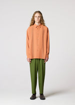 A person with long curly hair stands facing forward, wearing IM MEN's AIR PLEATS peach button-up shirt with vertical pleats and relaxed-fit olive green pants, paired with dark shoes, against a plain light gray background.