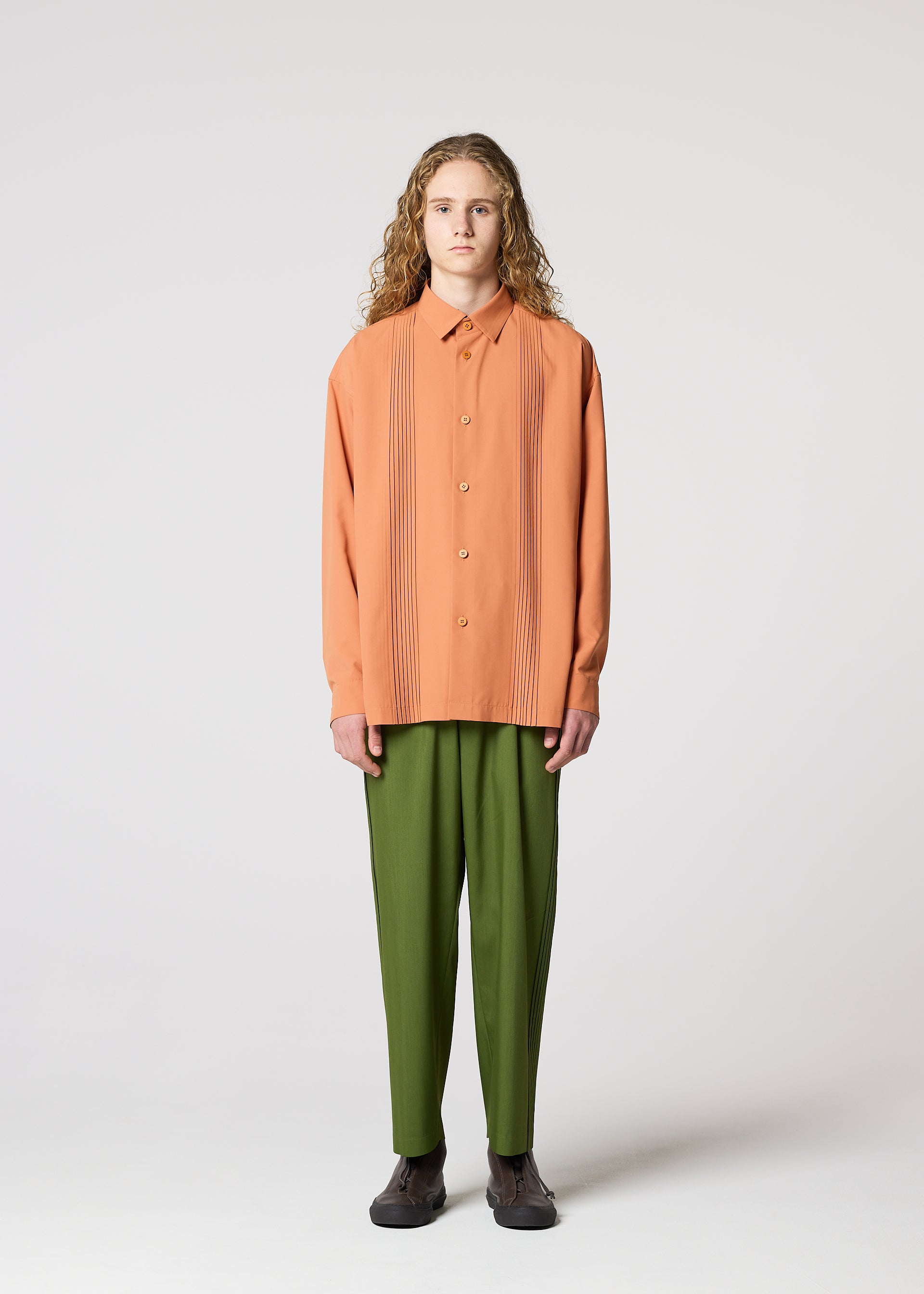 Zoomed view A person with long curly hair stands facing forward, wearing IM MEN's AIR PLEATS peach button-up shirt with vertical pleats and relaxed-fit olive green pants, paired with dark shoes, against a plain light gray background.