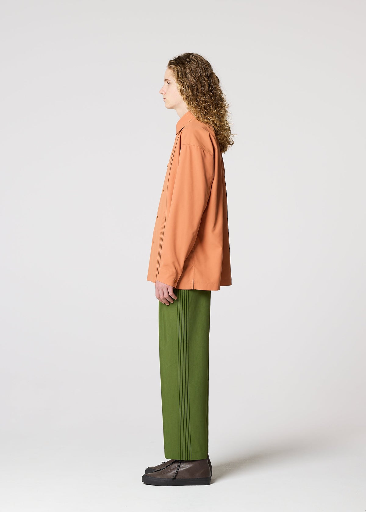 A person with long curly hair stands in profile against a plain white background, wearing IM MEN's AIR PLEATS relaxed fit green trousers, an orange button-up shirt, and dark shoes. view 3 of 5