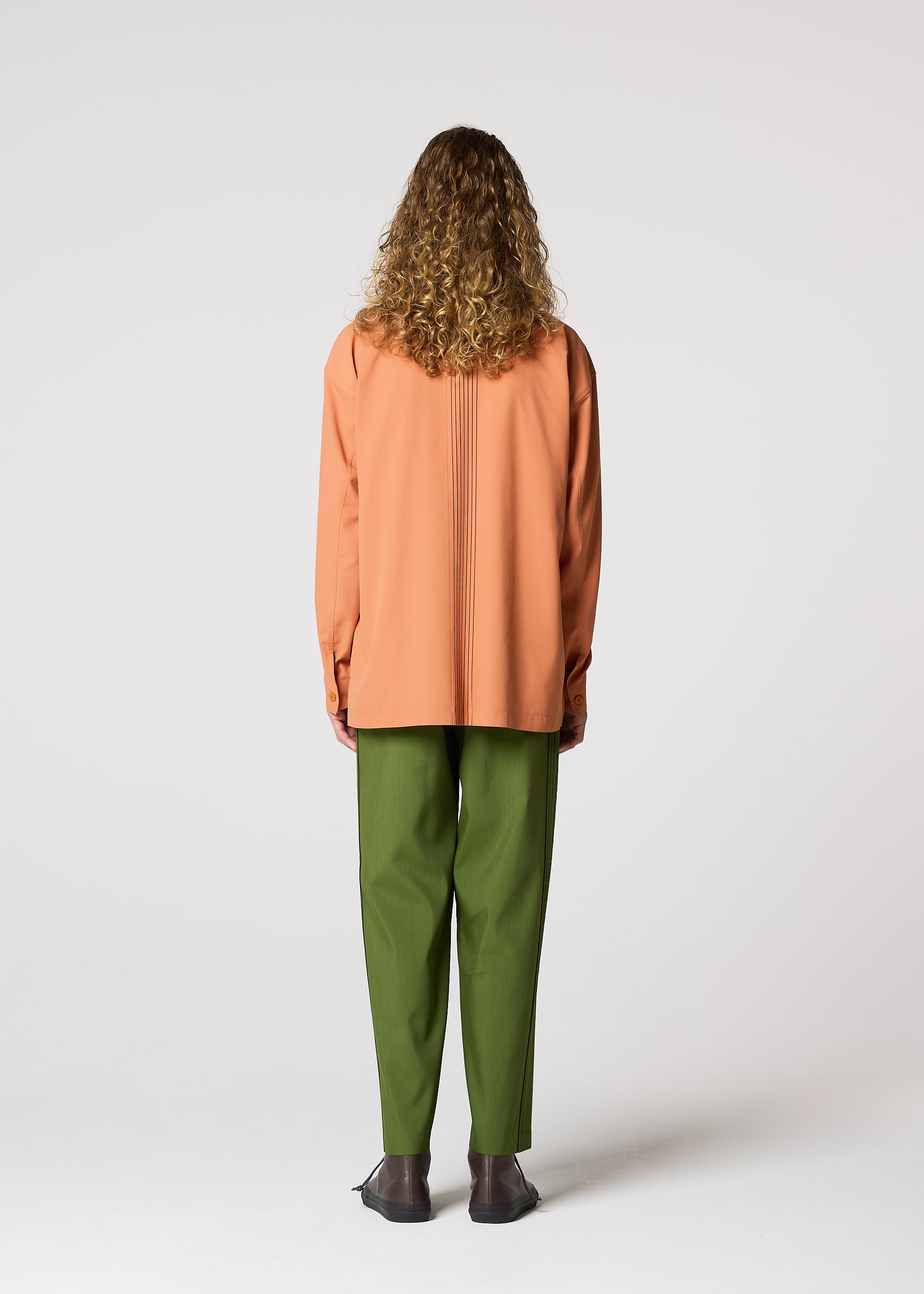 Zoomed view A person with curly hair, facing away, wears IM MEN's AIR PLEATS: an oversized orange shirt and relaxed-fit green pants with black shoes, standing against a plain white background.