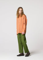A person with long curly hair wears IM MEN's AIR PLEATS orange button-up shirt and relaxed green pants with dark shoes, standing against a plain light gray background.