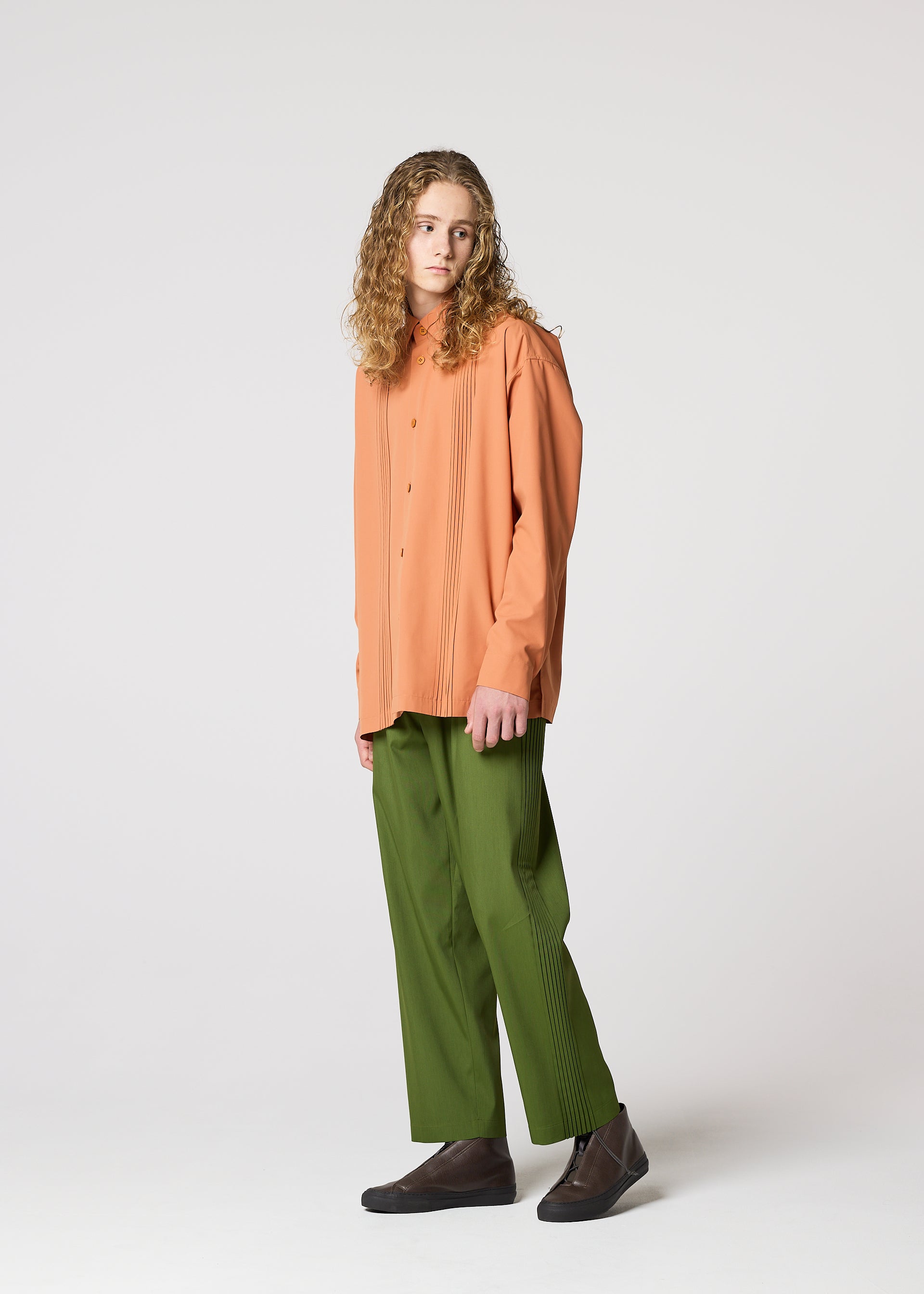 Zoomed view A person with long curly hair wears IM MEN's AIR PLEATS orange button-up shirt and relaxed green pants with dark shoes, standing against a plain light gray background.