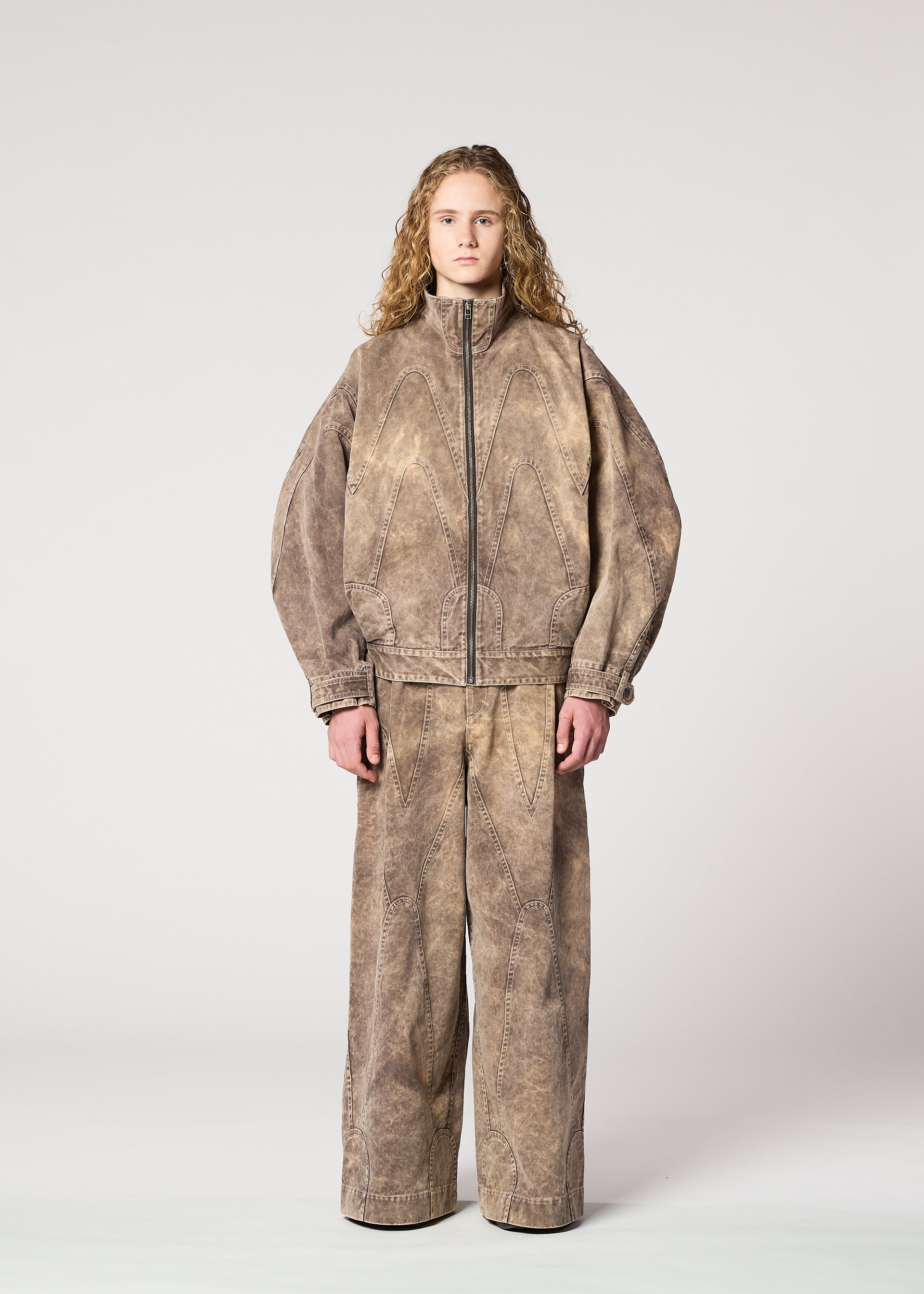 Zoomed view A person with long curly hair wears the IM MEN ELEMENTS loose-fitting brown textured jacket and matching wide pants, crafted from thick cotton, standing against a plain light gray background.