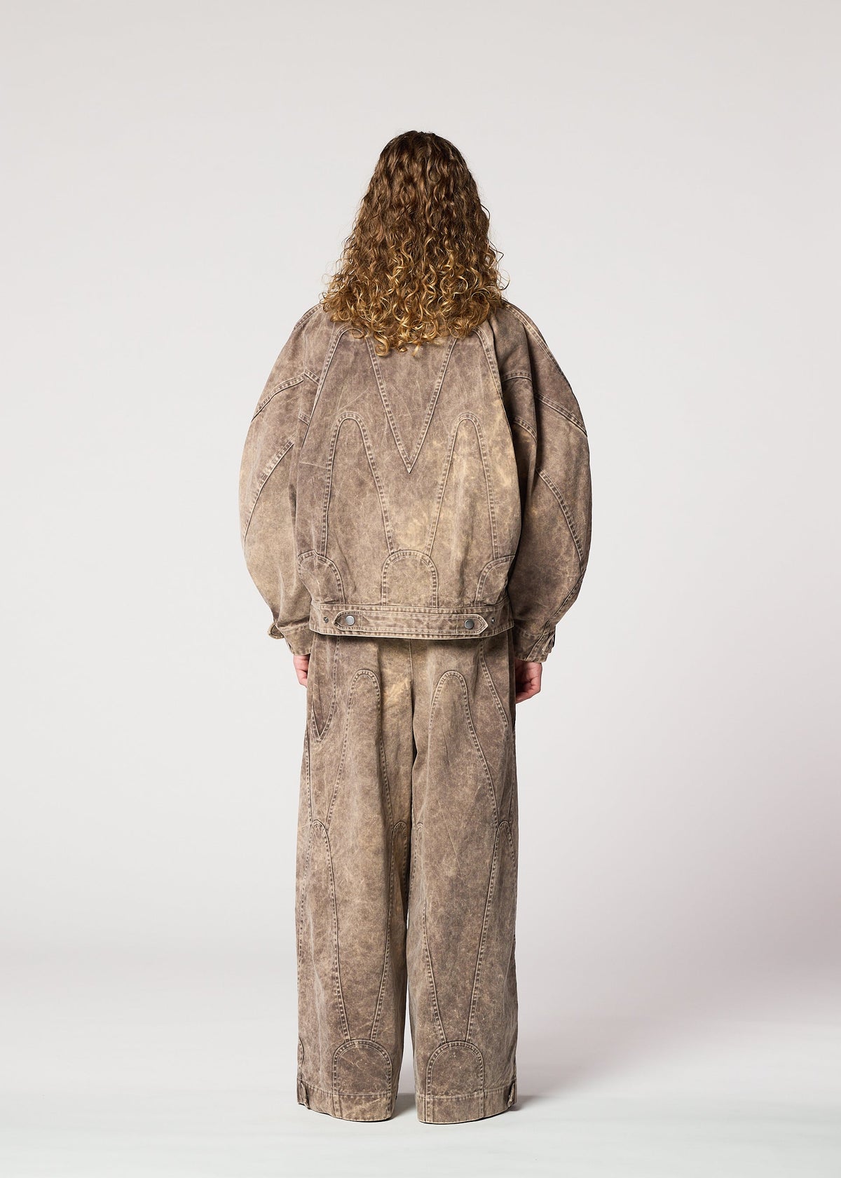 A person with curly hair faces away, wearing the IM MEN ELEMENTS set: an oversized taupe denim jacket and matching wide pants with visible seams, crafted from thick cotton, shown against a plain light background. view 10 of 11