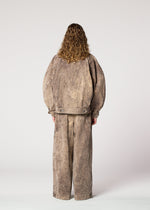 A person with curly hair faces away, wearing the IM MEN ELEMENTS set: an oversized taupe denim jacket and matching wide pants with visible seams, crafted from thick cotton, shown against a plain light background.