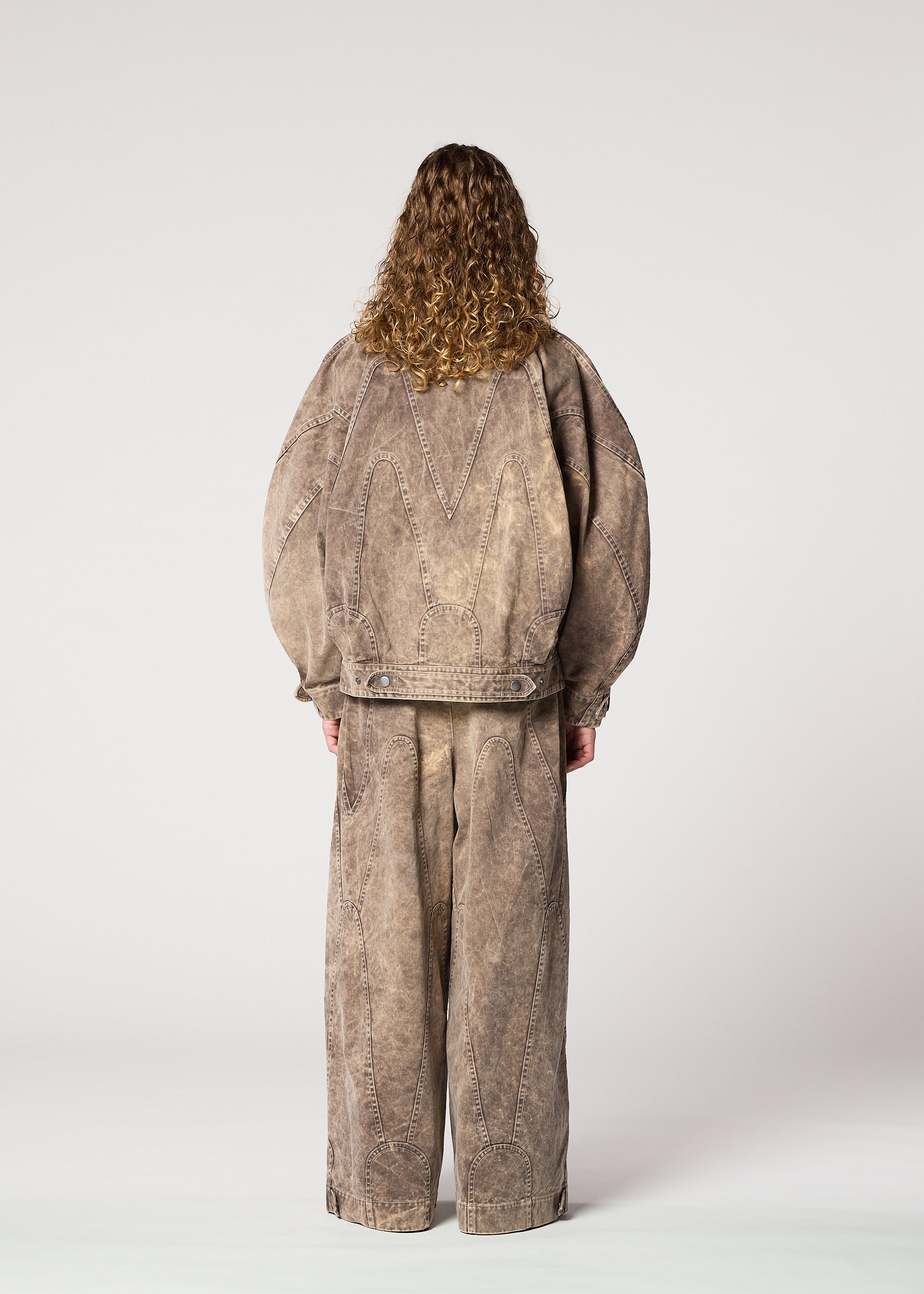 Zoomed view A person with curly hair faces away, wearing the IM MEN ELEMENTS set: an oversized taupe denim jacket and matching wide pants with visible seams, crafted from thick cotton, shown against a plain light background.