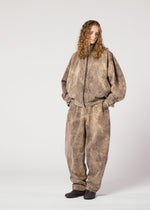 A person with long curly hair stands against a plain background, wearing the ELEMENTS oversized brown jacket and wide-legged pants by IM MEN, crafted from thick cotton with a subtle color-fading effect. They look to the side with hands in pockets.