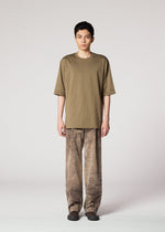 A person stands facing forward in IM MEN’s ELEMENTS olive green T-shirt and ceramic artist-inspired pants with a subtle color-fading effect, paired with dark shoes, set against a plain light grey background.