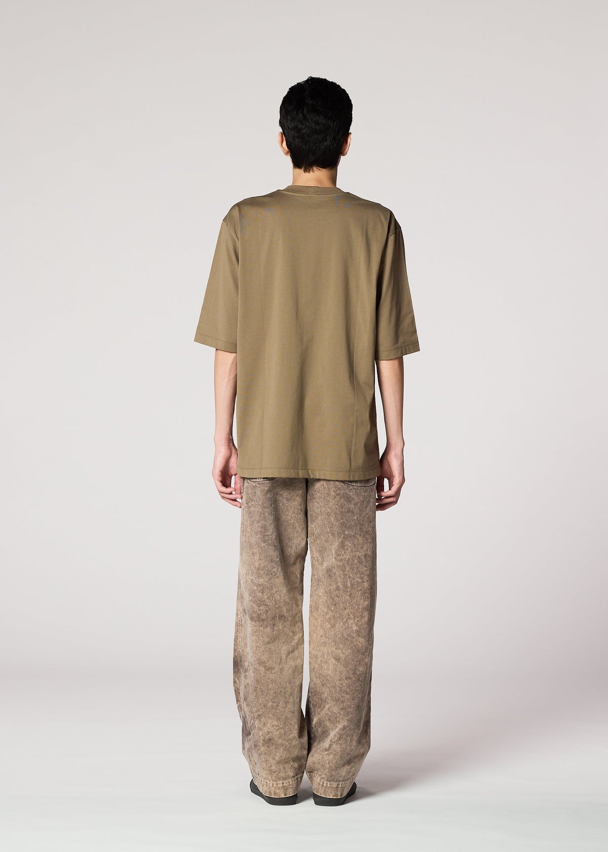 A person stands facing away from the camera, wearing IM MEN's ELEMENTS olive-green tee and ceramic artist-inspired thick cotton pants with a subtle faded effect, set against a plain light background. view 7 of 8