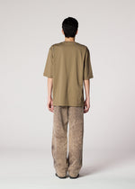 A person stands facing away from the camera, wearing IM MEN's ELEMENTS olive-green tee and ceramic artist-inspired thick cotton pants with a subtle faded effect, set against a plain light background.