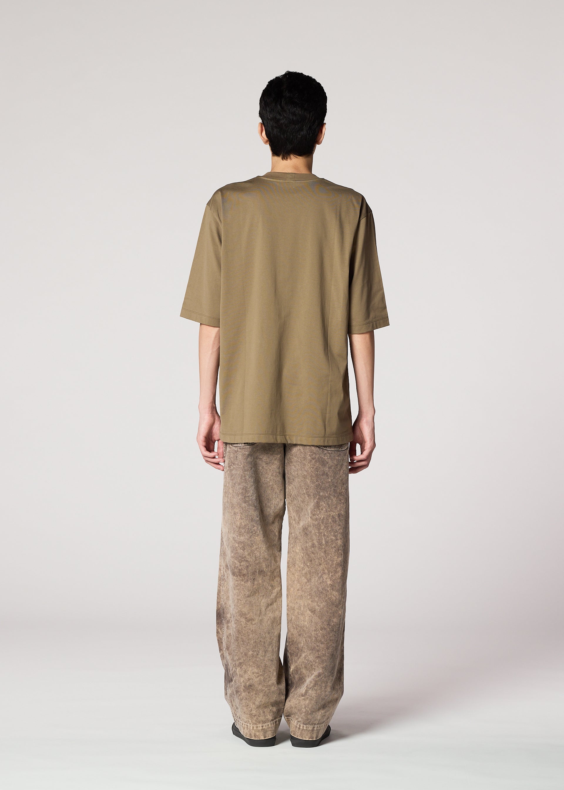 Zoomed view A person stands facing away from the camera, wearing IM MEN's ELEMENTS olive-green tee and ceramic artist-inspired thick cotton pants with a subtle faded effect, set against a plain light background.