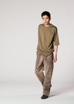 A person with short dark hair wears IM MEN's ELEMENTS: a loose olive t-shirt and wide-legged, faded brown cotton pants, standing against a plain light background.