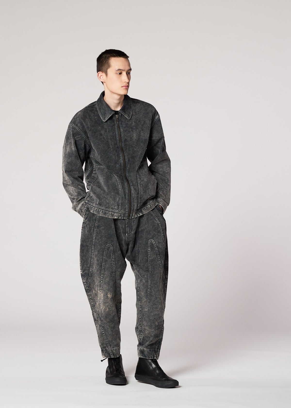 A person wears the IM MEN ELEMENTS dark, washed-out denim jacket and irregular-cut pants with a unique color-fading effect, paired with black ankle boots, standing against a light gray background with hands in pockets. view 8 of 8