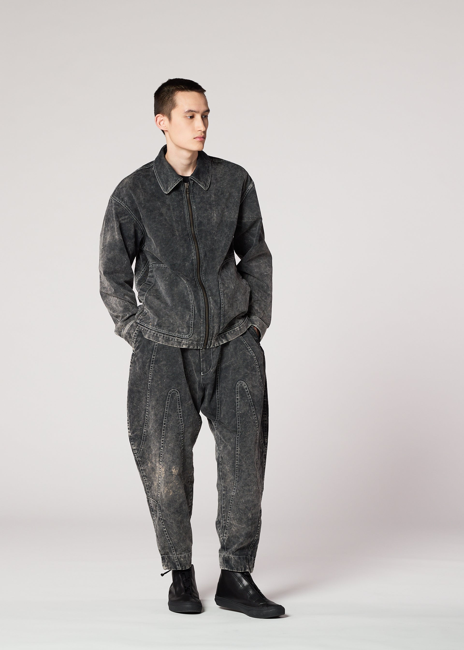 Zoomed view A person wears the IM MEN ELEMENTS dark, washed-out denim jacket and irregular-cut pants with a unique color-fading effect, paired with black ankle boots, standing against a light gray background with hands in pockets.