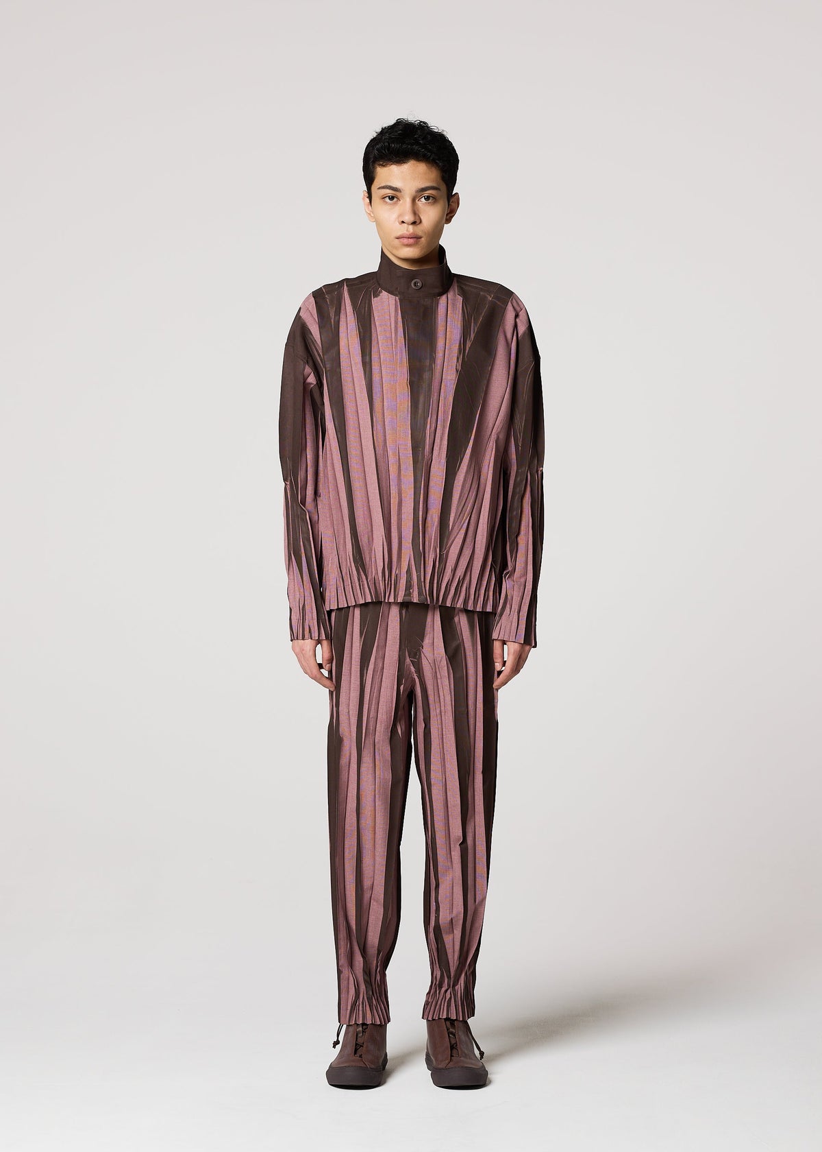 A person faces forward wearing IM MEN’s EDGE, a loose-fitting set with vertical brown and mauve stripes, long sleeves, elastic cuffs, and Shoji Kamoda ceramic inspired pants, against a plain light background. view 6 of 9