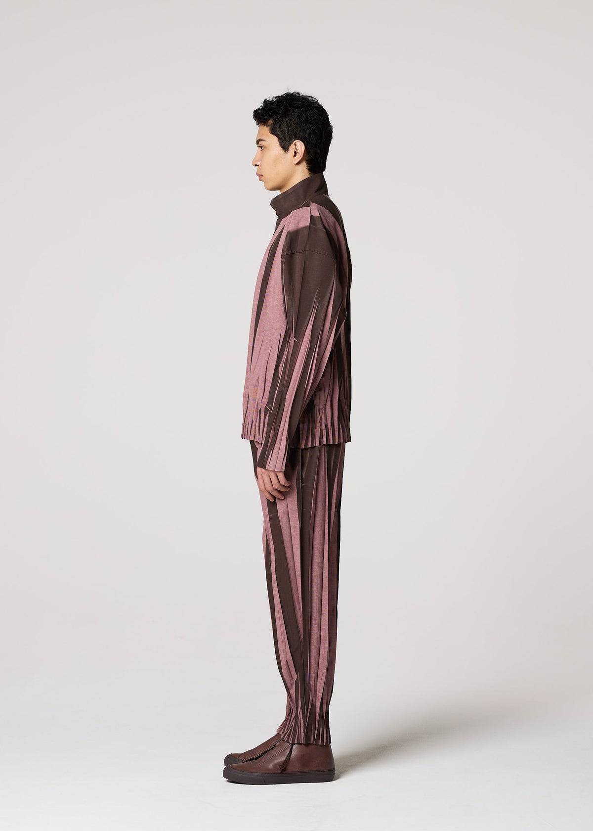 A person stands in profile facing left, dressed in the IM MEN EDGE suit with brown and mauve ceramic-inspired striped pants and brown shoes, set against a light gray studio background. view 7 of 9