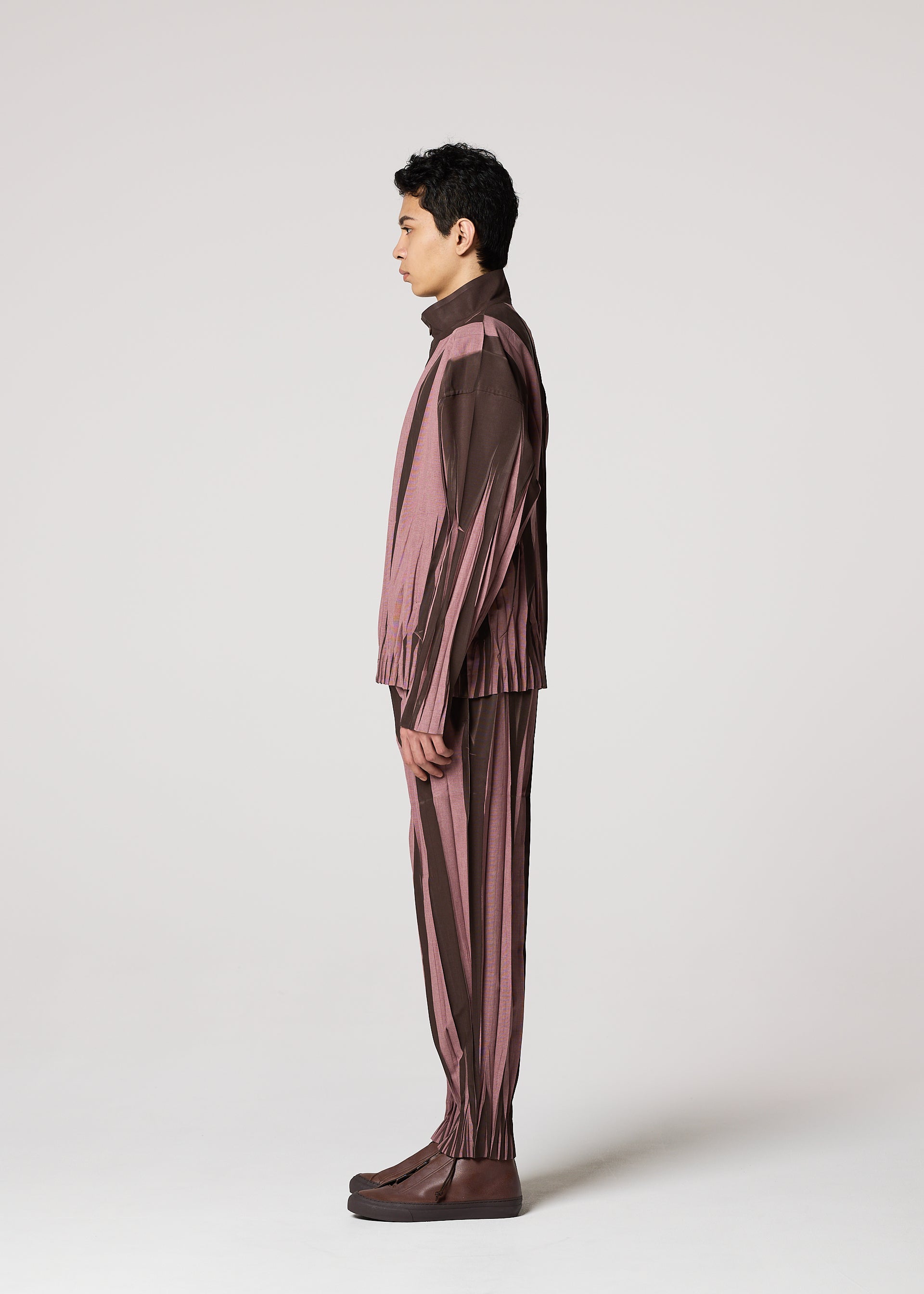 Zoomed view A person stands in profile facing left, dressed in the IM MEN EDGE suit with brown and mauve ceramic-inspired striped pants and brown shoes, set against a light gray studio background.